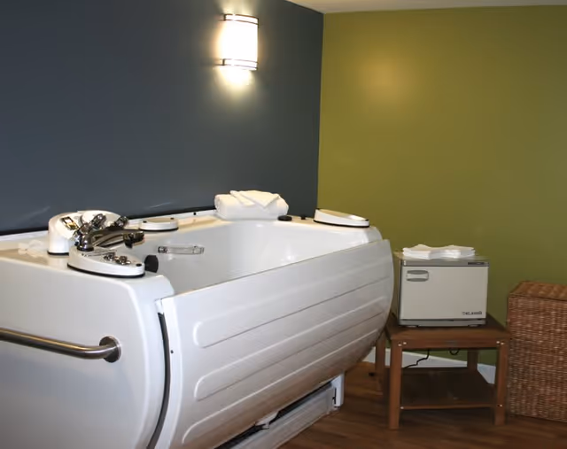 A white therapeutic bathtub with control knobs and a handrail on the side, placed against a wall painted half dark gray and half green. A small wooden table next to the tub holds a small white appliance and folded towels. A wicker basket is also visible beside the table. The floor is wooden, and a wall-mounted light fixture is above the tub.