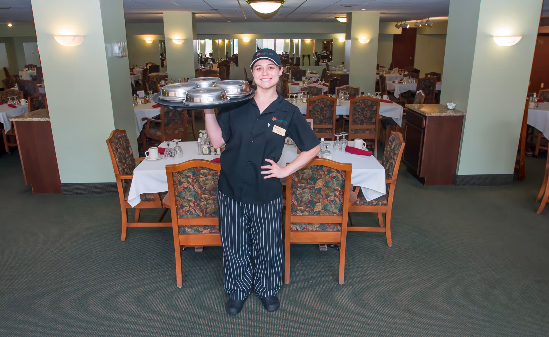 A smiling server wearing a black uniform and striped pants stands in a dining room holding a tray with covered dishes. The dining room has multiple tables set with white tablecloths, glasses, and napkins, and floral upholstered chairs. The room is well-lit with wall sconces and ceiling lights.