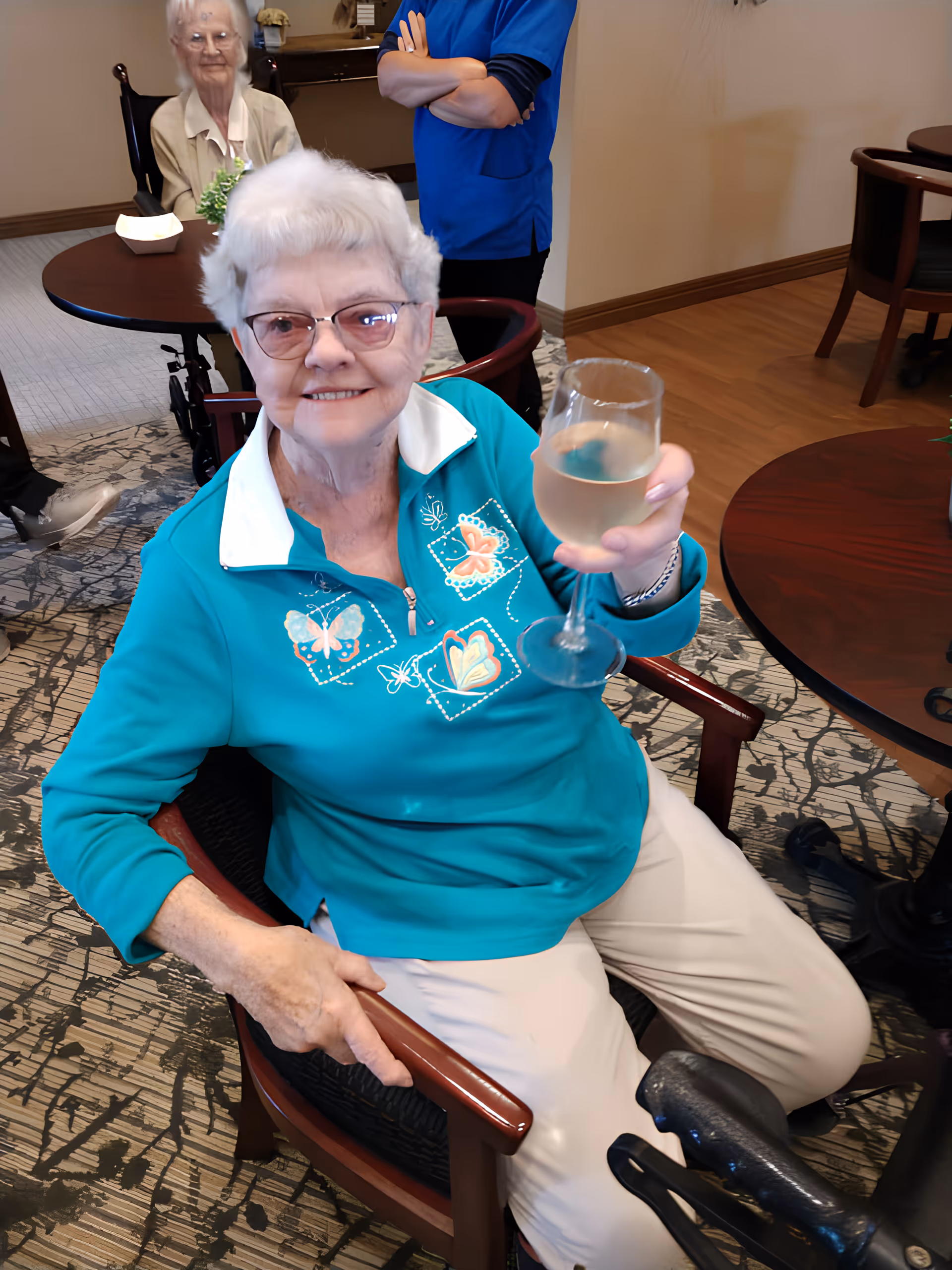 An elderly woman with short white hair and glasses is sitting in a chair, smiling and holding up a glass of white wine in a toast. She is wearing a teal shirt with butterfly designs and light-colored pants. In the background, another elderly woman is seated at a round table, and a person in a blue uniform stands with arms crossed. The setting appears to be a comfortable indoor common area with patterned carpet and wooden furniture.