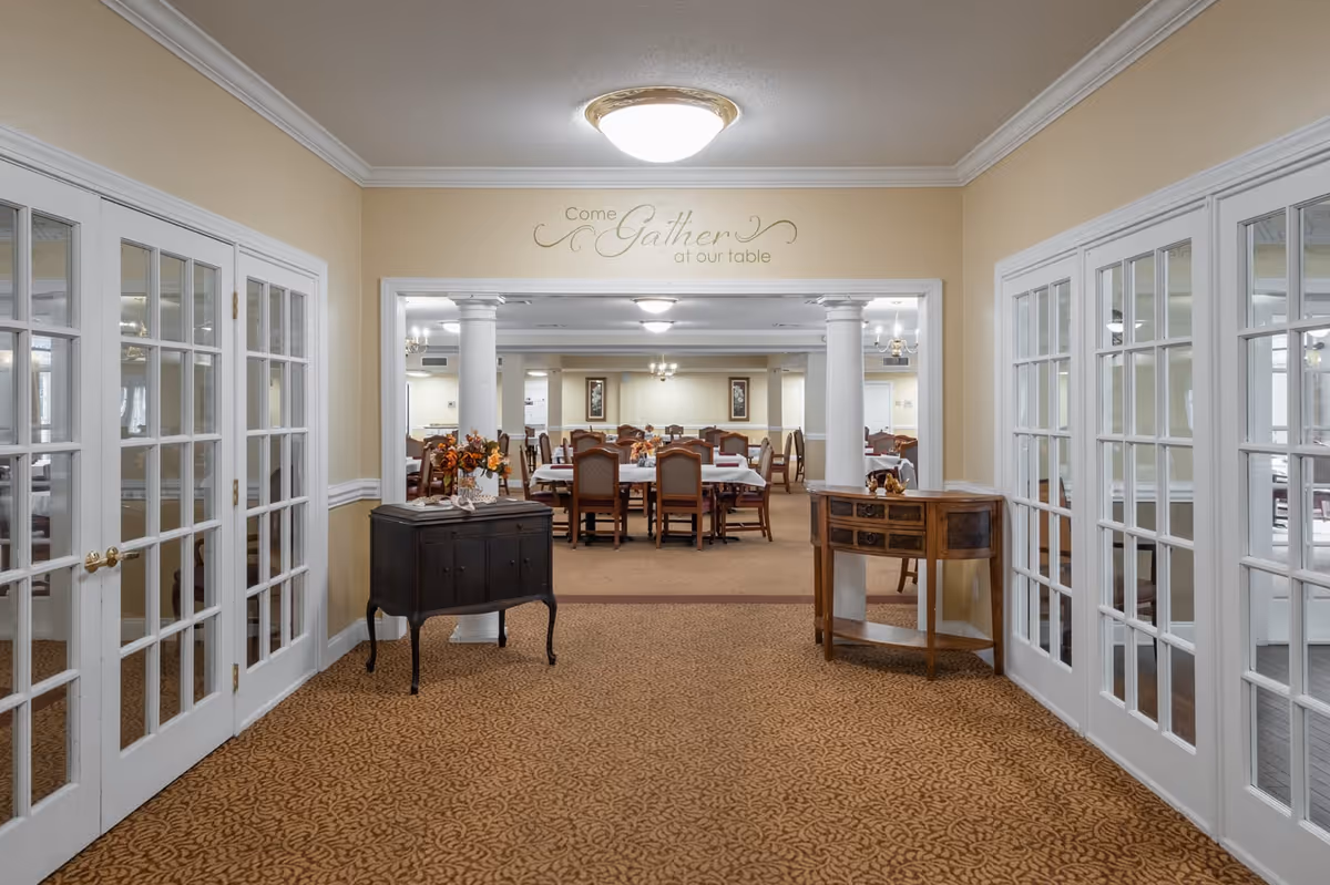 View through a hallway with double glass doors on both sides leading to a dining area with tables and chairs. The walls are beige with white trim, and there are two small wooden tables with decorative items on either side of the hallway. Above the entrance to the dining area, a wall decal reads 'Come Gather at our table'.