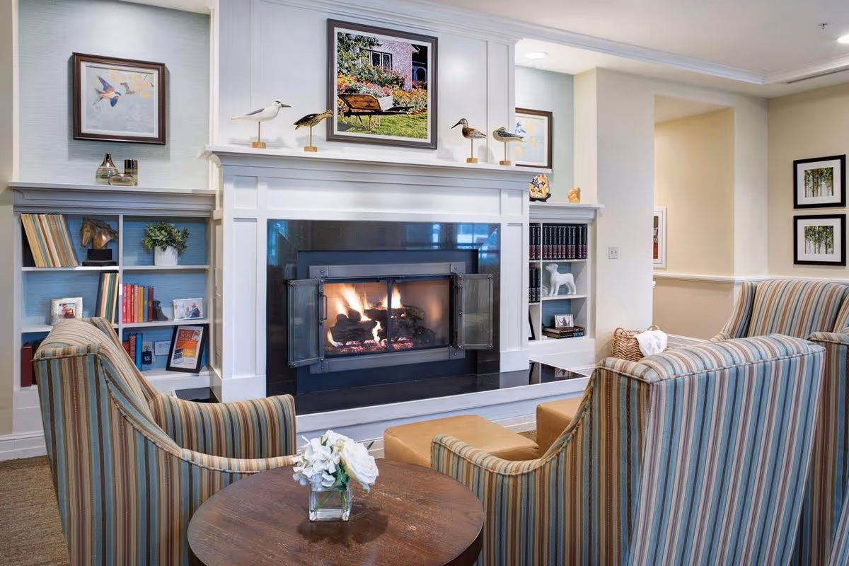 Cozy living room area with a lit fireplace surrounded by white mantel and black tiles. Two striped armchairs face the fireplace with a small round wooden table between them holding a small vase with white flowers. Built-in shelves on either side of the fireplace hold books, framed photos, and decorative items. Artwork and bird figurines decorate the mantel and walls.