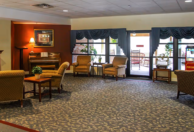 A spacious common area with patterned carpet, several upholstered armchairs arranged around small wooden tables with potted plants, a piano with a lamp and framed picture on the wall behind it, and large windows and a glass door leading to an outdoor patio with rocking chairs.