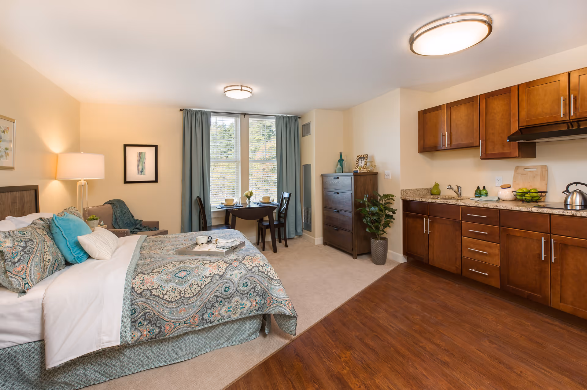 Furnished studio apartment interior with a bed, bedside lamp, small dining table by a window, dresser, and a kitchenette with wooden cabinets.