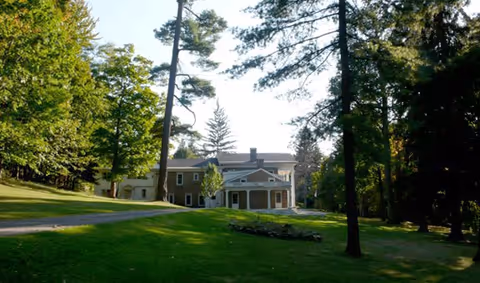 A large, two-story building surrounded by tall trees and green grass under a clear sky, with a paved driveway leading up to the entrance.