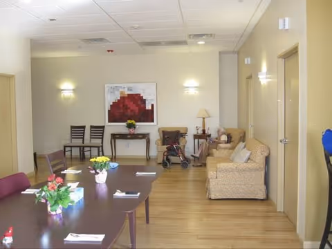 Communal room with a long dining table and chairs, armchairs, a walker, and wall art in an assisted living facility.