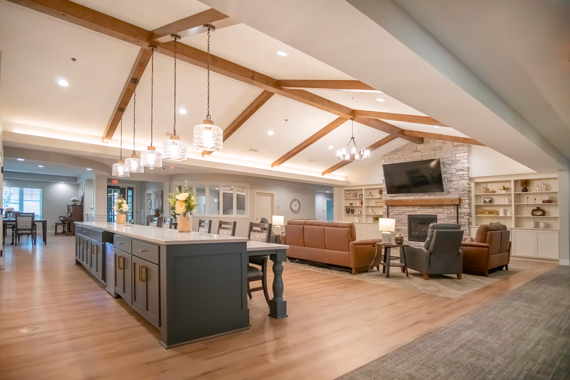 Spacious open interior showing a communal living area with a large kitchen island, pendant lights, seating area with sofas and armchairs, and a stone fireplace with TV under vaulted ceilings and exposed wooden beams.