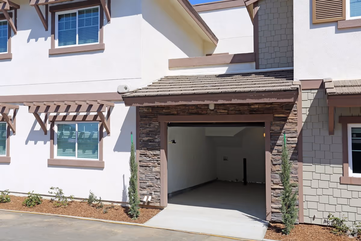 Exterior view of a residential building showing a garage with an open door, stone and shingle siding, white walls with brown trim, two windows with shutters, and small landscaped plants along the building.