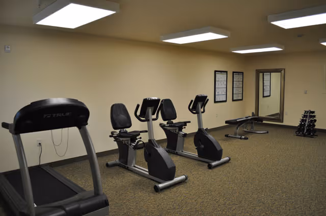 A small fitness room with a treadmill, two recumbent exercise bikes, a weight bench, a dumbbell rack, and a wall mirror under fluorescent ceiling lights.