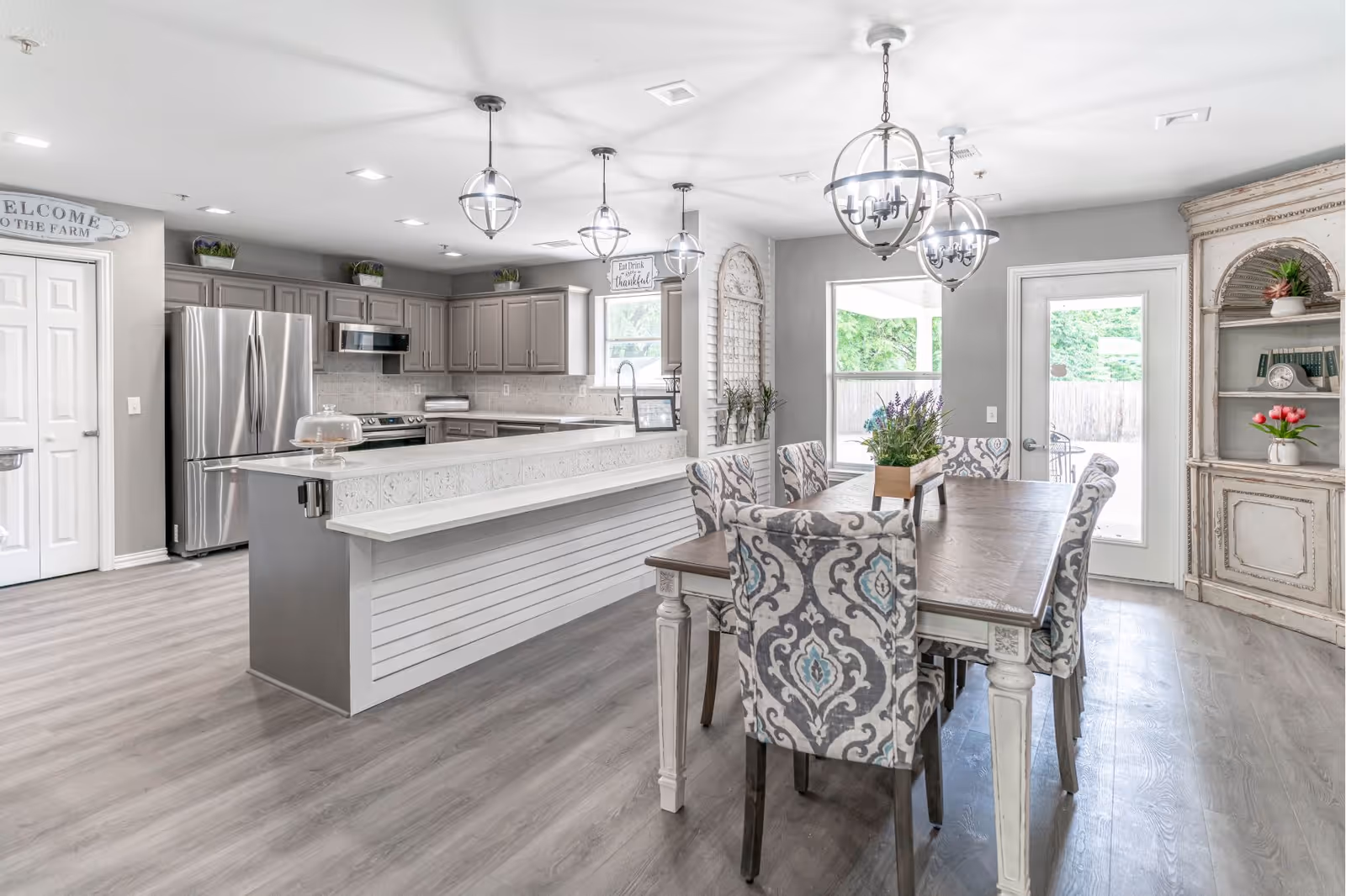 Bright and spacious kitchen and dining area with gray cabinetry, stainless steel refrigerator and oven, a long white island with decorative paneling, and a wooden dining table surrounded by patterned upholstered chairs. The room features multiple hanging light fixtures, large windows, a glass door leading outside, and a vintage-style cabinet with plants and books.