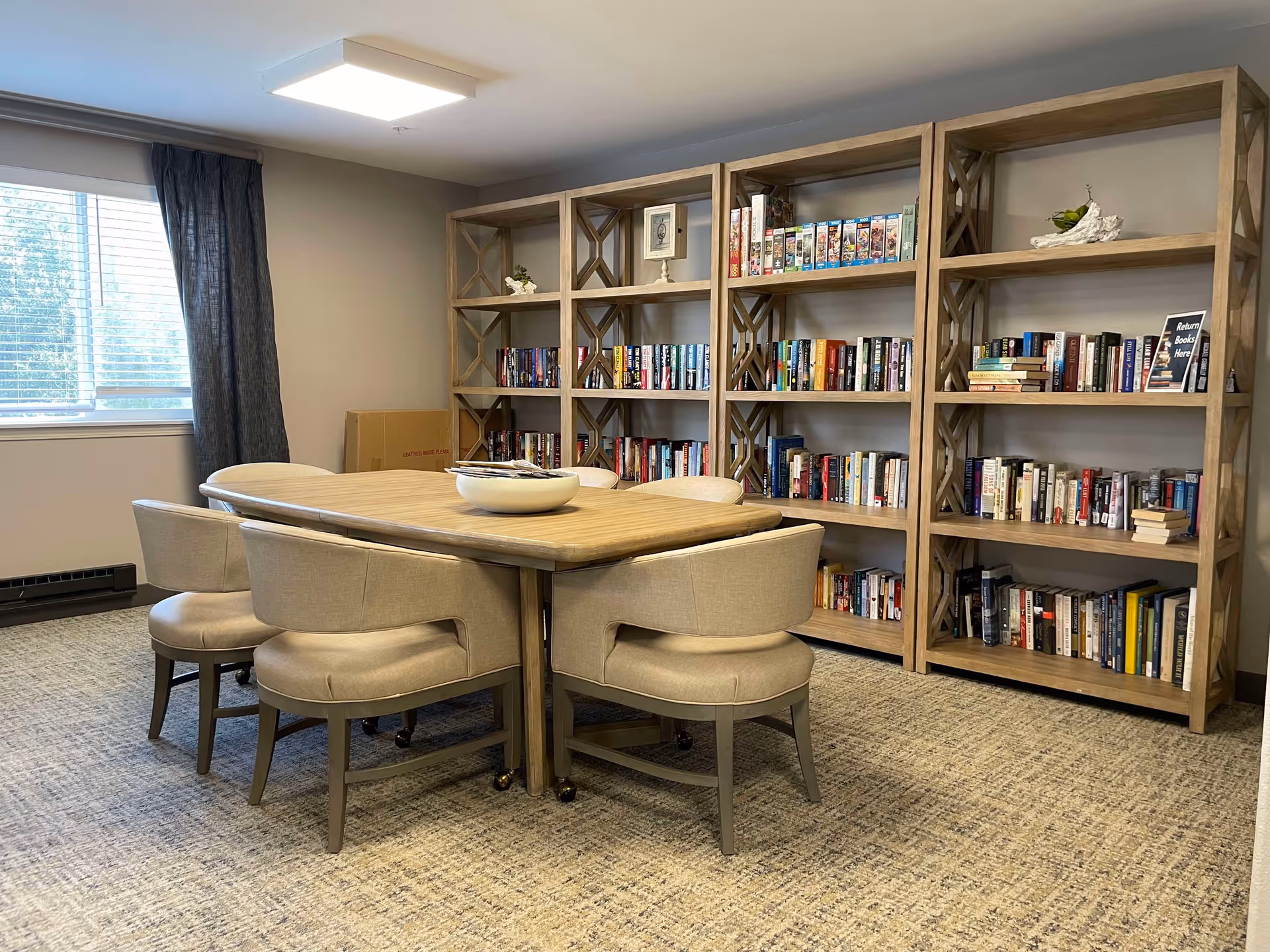 A cozy room with a wooden table surrounded by six beige upholstered chairs. Behind the table are three large wooden bookshelves filled with various books. A window with dark curtains allows natural light into the room, and the floor is carpeted in a neutral tone.