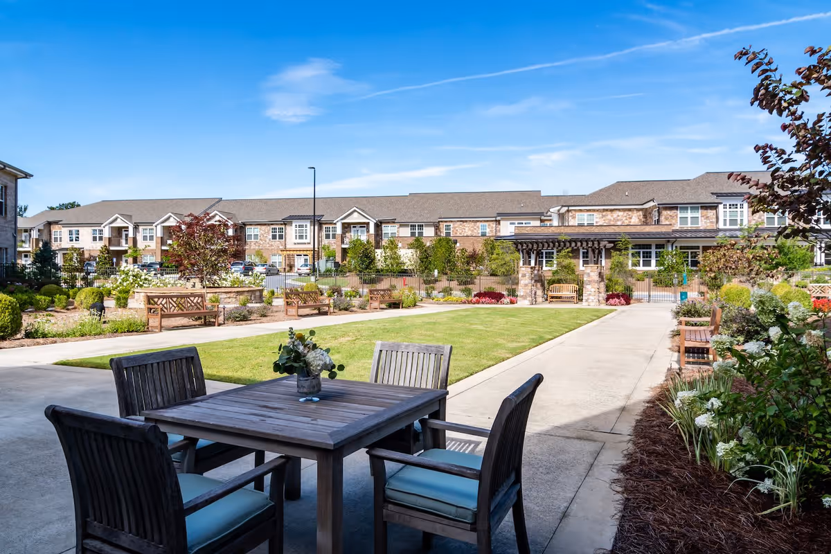 Outdoor courtyard area at Arbor Terrace Hamilton Mill featuring a wooden table with four cushioned chairs, a grassy lawn, benches, landscaped garden beds, and a pergola structure with a bench underneath. The background shows a two-story brick building under a clear blue sky.