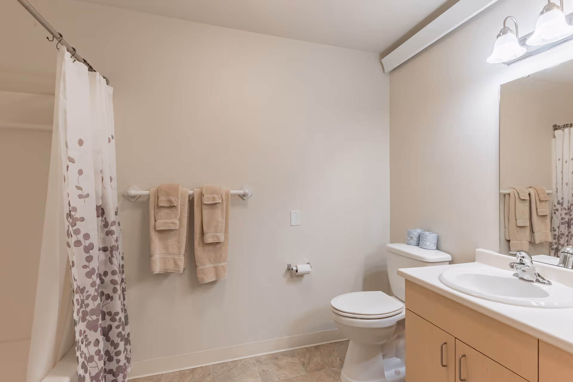 A clean bathroom with a toilet, sink and vanity, shower with a patterned curtain, and towels hanging on a rack.