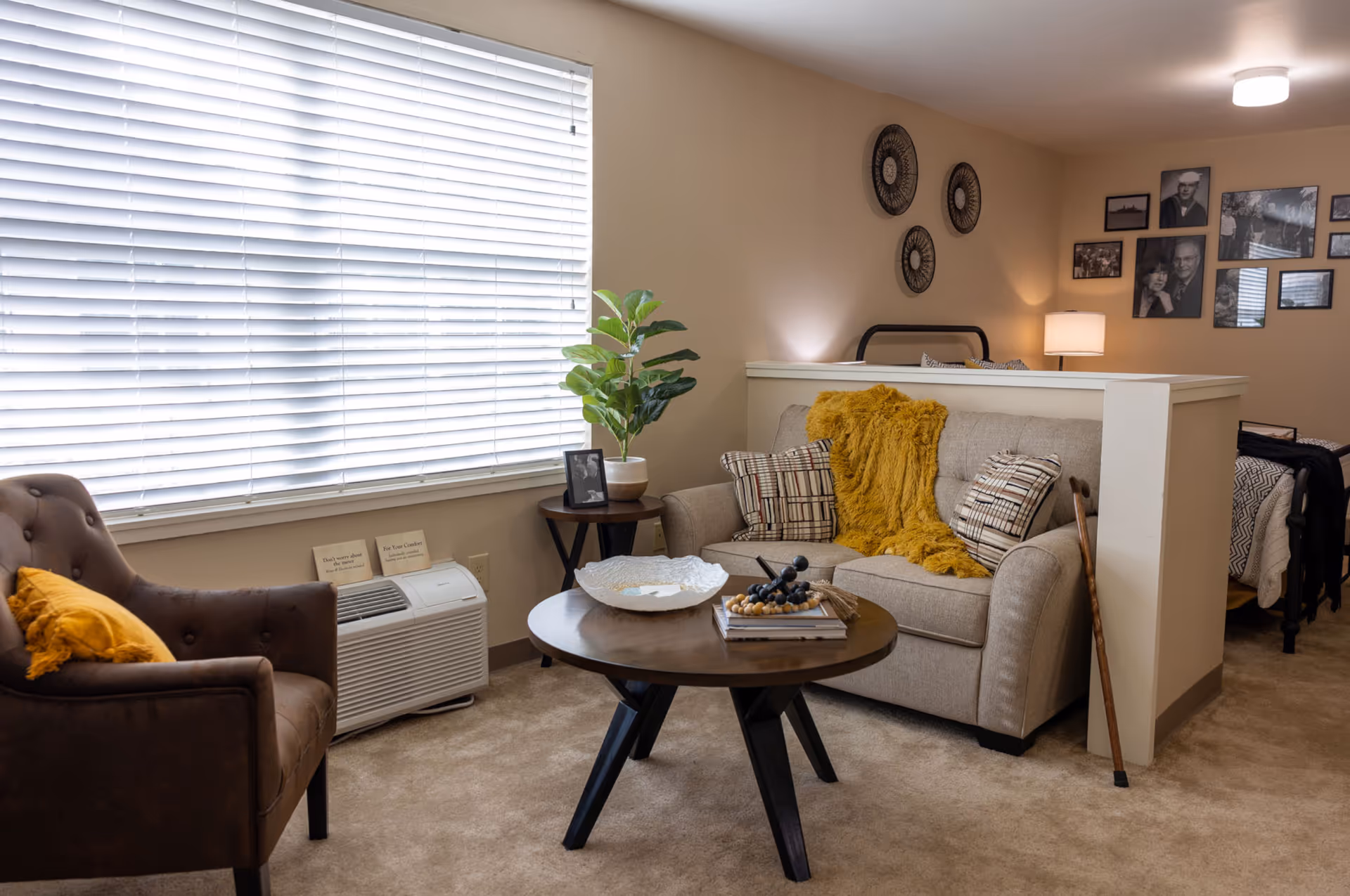 A cozy living area in a senior living facility featuring a beige loveseat with patterned pillows and a mustard yellow throw blanket, a round wooden coffee table with decorative items, a brown armchair with a yellow pillow, a small side table with a potted plant and framed photo, and a walking cane leaning against a half wall. Behind the half wall is a bed with black metal frame and black and white bedding. The walls are decorated with black and white photos and circular wall art. A large window with white blinds lets in natural light.