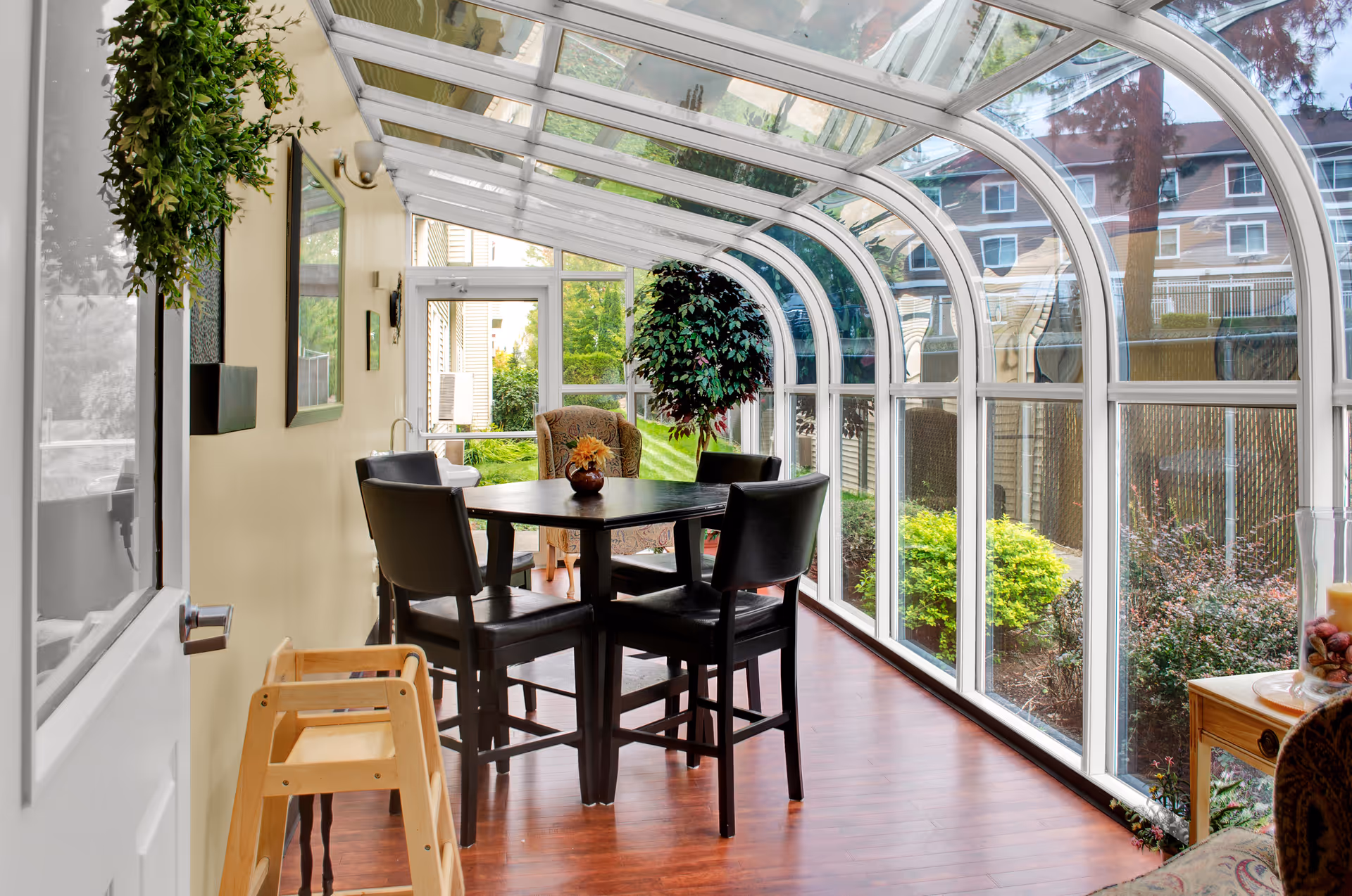 A bright sunroom with a curved glass ceiling and walls, featuring a dark wooden table surrounded by four black chairs. There is a potted plant on the table and a large leafy plant near the far end. The floor is wooden, and outside the glass walls, green bushes and a building are visible.