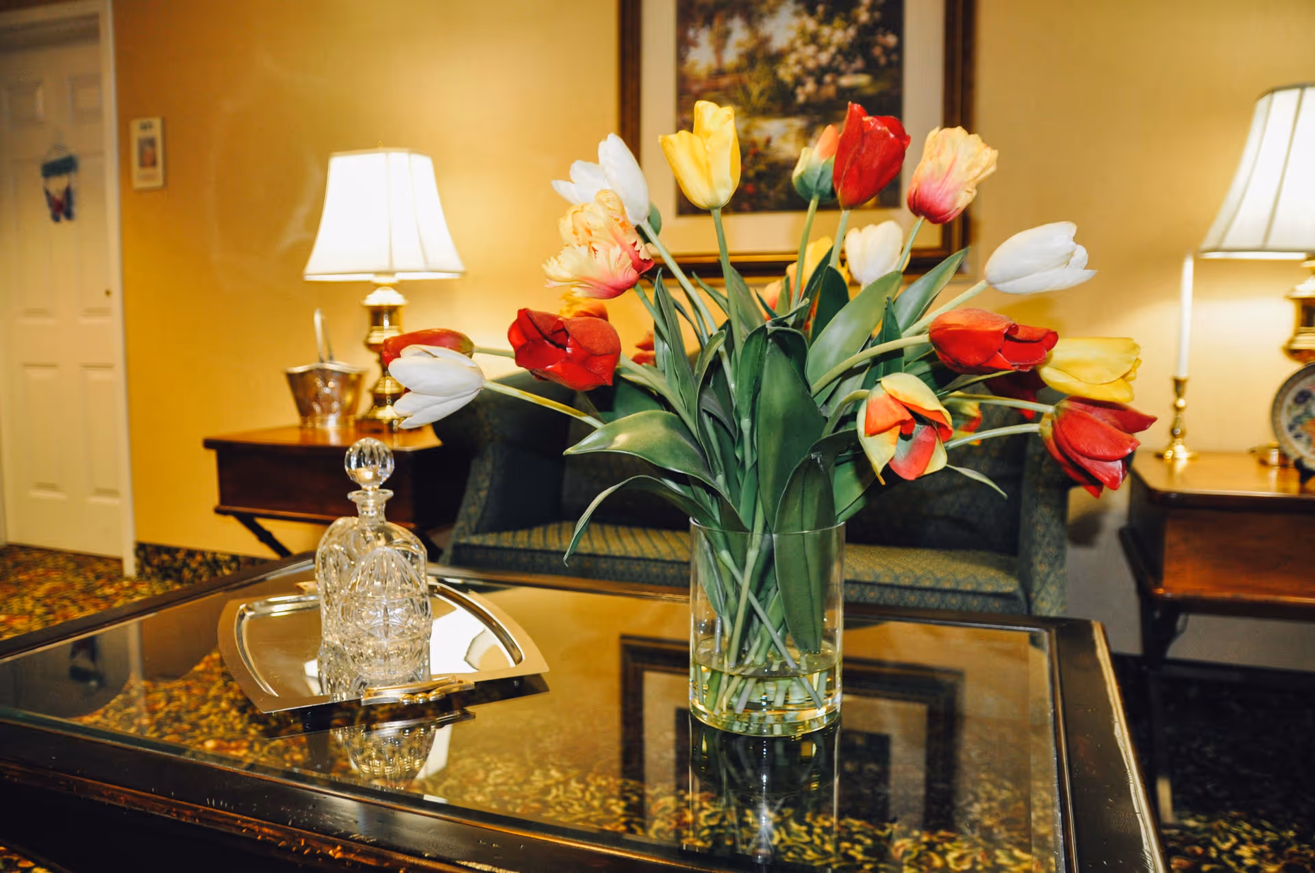 A glass vase filled with colorful tulips in red, yellow, white, and pink sits on a reflective glass coffee table. Behind the table, there is a green upholstered sofa, two wooden side tables with brass lamps, and a framed floral painting on a beige wall. The room has a warm, cozy ambiance with patterned carpet and soft lighting.