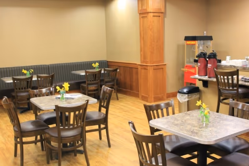 A dining area with several tables and wooden chairs arranged on a wooden floor. Each table has a small vase with yellow flowers. There is a popcorn machine and two beverage dispensers on a counter in the background, along with a trash can underneath the counter. The walls are painted beige with wooden paneling and trim.