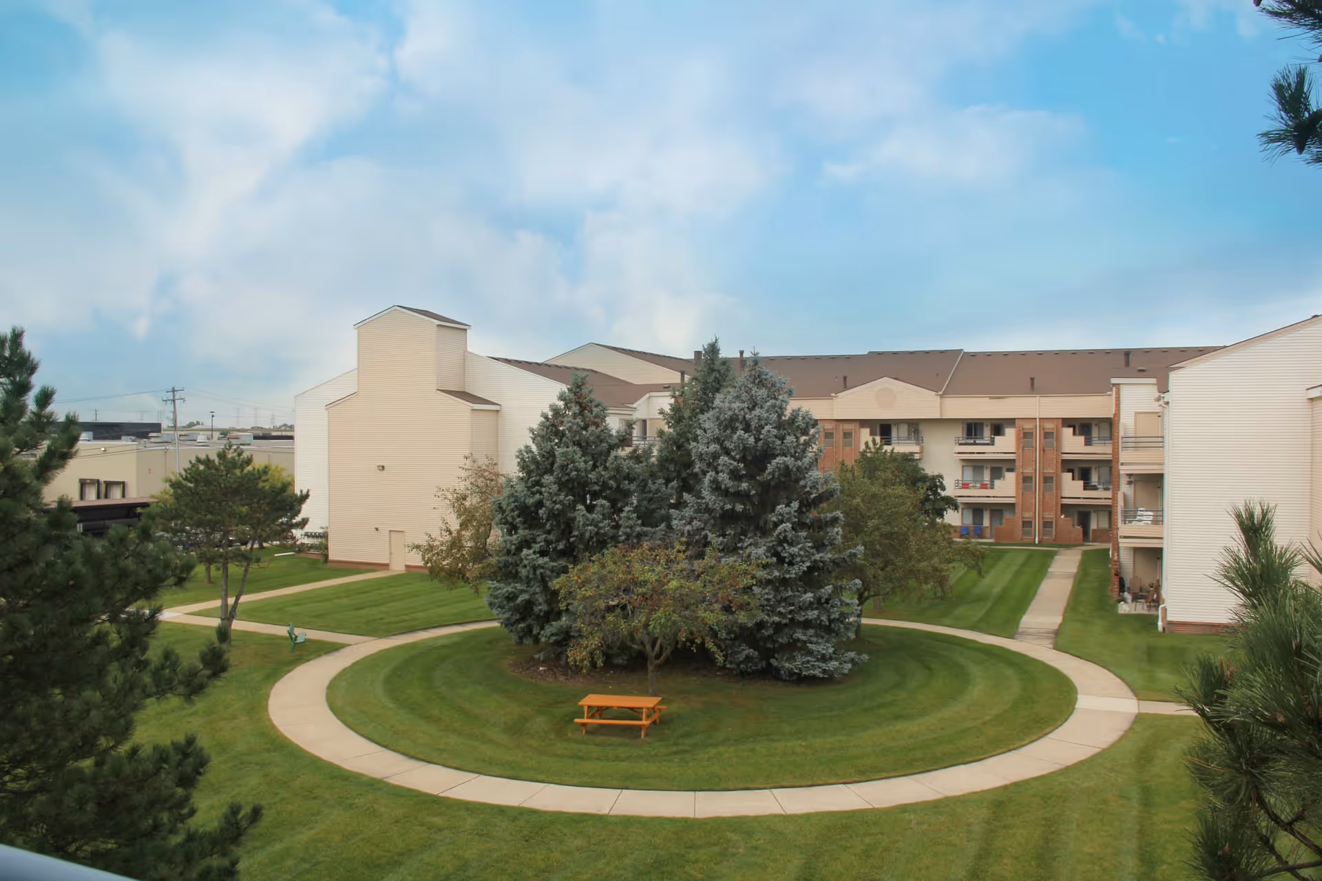 Outdoor courtyard area of Shelby Manor Senior Living featuring a circular walkway surrounding a cluster of trees and a wooden picnic table, with multi-story residential buildings in the background under a partly cloudy sky.