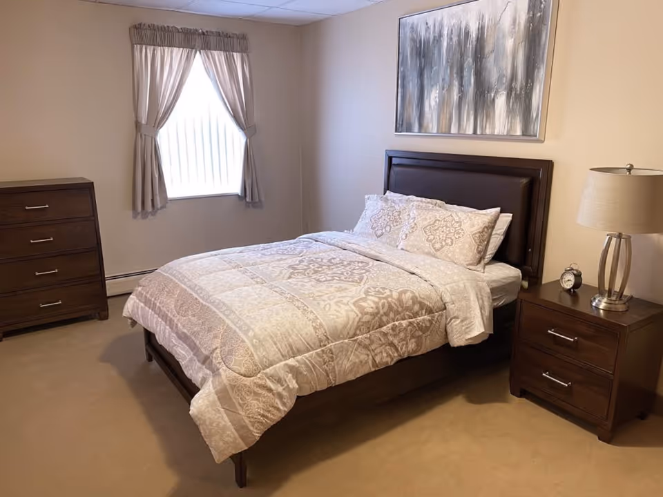 A neatly made bed with patterned bedding in a simple bedroom featuring a wooden headboard, matching nightstand and dresser, a lamp, a window with curtains, and abstract wall art.