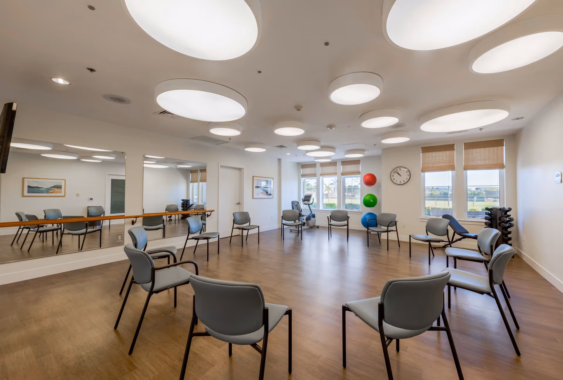 A spacious exercise or activity room with a circle of gray chairs on a wooden floor. The room has large round ceiling lights, a wall clock, three large windows with blinds, a mirror along one wall, and exercise equipment including colorful exercise balls and a rack of weights.