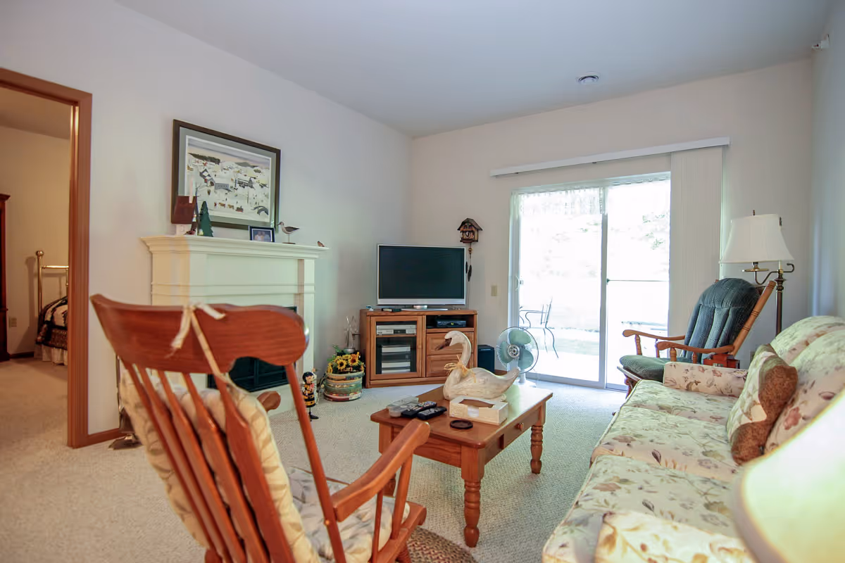 A cozy living room with a floral-patterned sofa, wooden rocking chair, and a wooden coffee table with decorative items. A TV sits on a wooden stand against the wall, next to a sliding glass door that leads outside. A fireplace with a framed picture above it is visible, and an adjacent doorway leads to a bedroom.