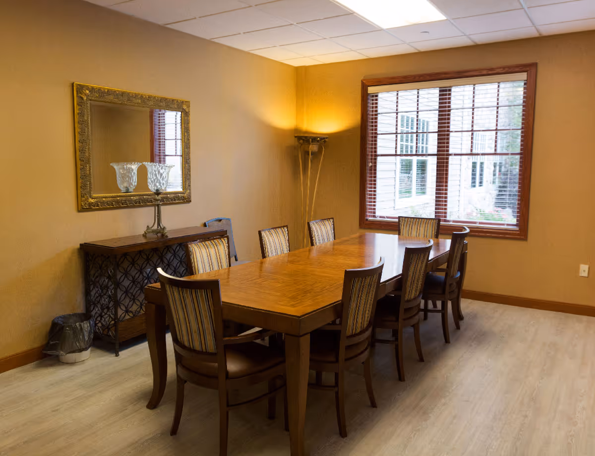A dining room with a long wooden table surrounded by chairs, a sideboard with a mirror, and a window with blinds.