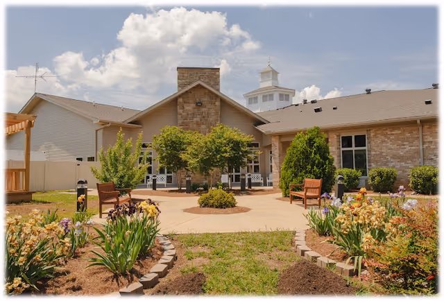 Outdoor garden area at The Pavilion Senior Living at Lebanon featuring a paved circular walkway with wooden benches, surrounded by flower beds and greenery, with a building in the background under a partly cloudy sky.
