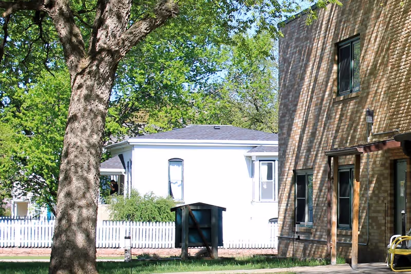 Outdoor view of a brick building beside a white house with a tree and lawn.