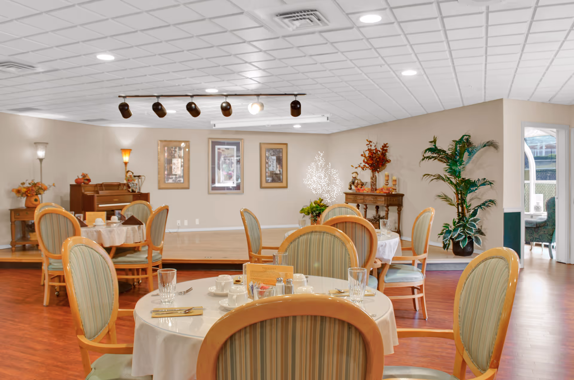 Bright dining room in a senior living facility with round tables set for meals, striped upholstered chairs, a small stage area with a piano, and decorative plants.