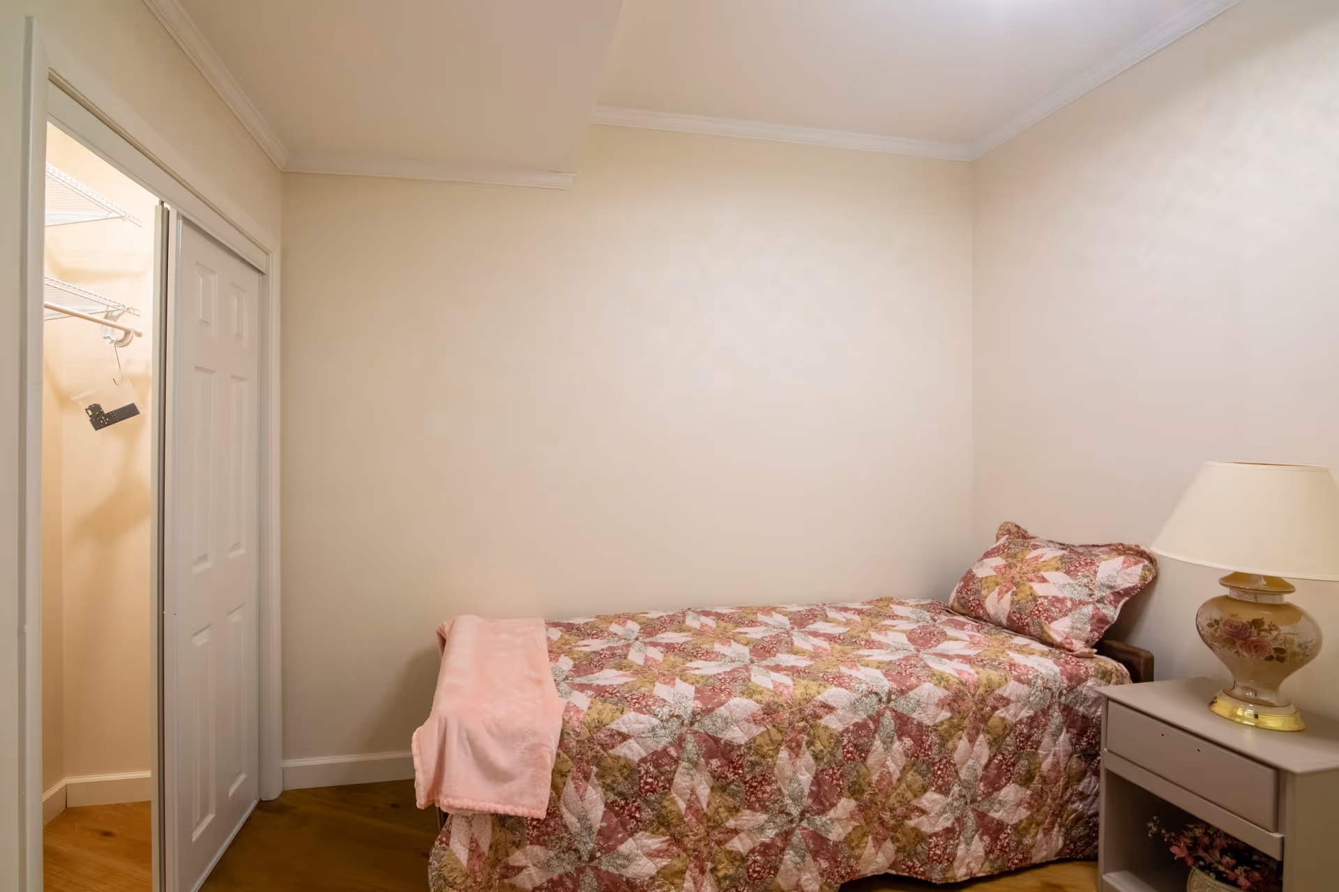 A small bedroom with a single bed covered in a patterned quilt with shades of pink, red, and green. There is a matching pillow and a pink blanket draped over the foot of the bed. Next to the bed is a nightstand with a floral lamp. On the left side of the room, there is an open closet with white sliding doors and a hanging rod with a few hangers.