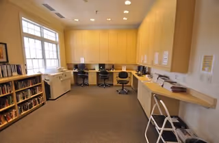A well-lit office or workroom with a large window on the left, built-in wooden cabinets and desks along the walls, three office chairs, a printer, bookshelves filled with books, and a folding chair in the foreground.