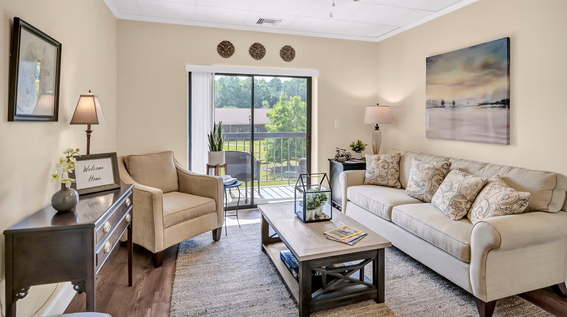 Bright furnished living room with a beige sofa, armchair, coffee table, lamps, and a sliding glass door opening to a balcony.