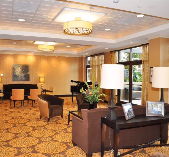A spacious and elegantly decorated living room area with patterned carpet, brown upholstered chairs and sofas, a black grand piano, and large windows with curtains letting in natural light. The room features warm lighting with ceiling fixtures and table lamps, a console table with framed photos and a flower arrangement, and a painting on the far wall.