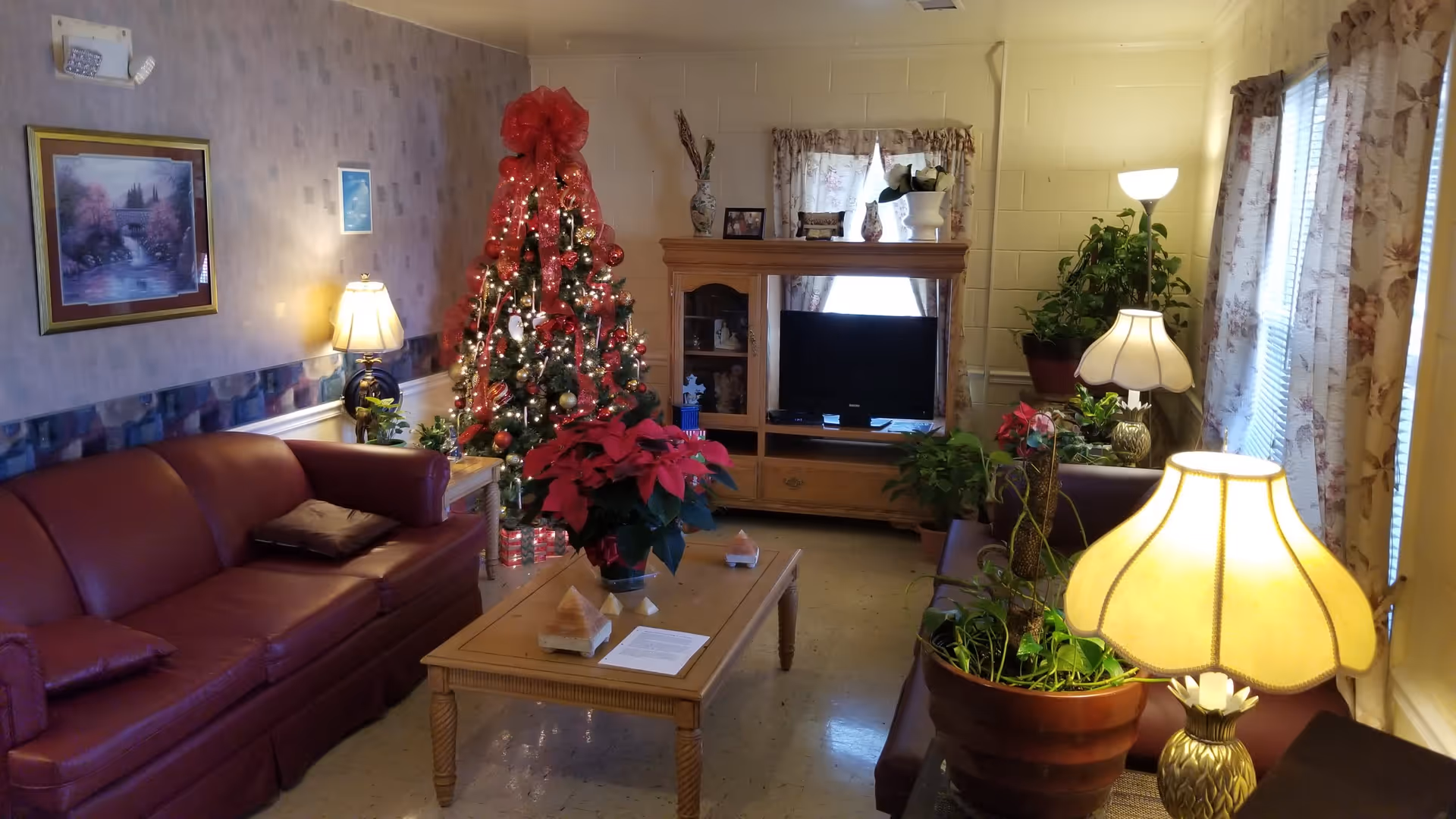 A cozy living room decorated for Christmas with a lit Christmas tree adorned with red ribbons and ornaments. The room features two maroon leather sofas, a wooden coffee table with a poinsettia plant and some papers on it, several table lamps, potted plants, a wooden entertainment center with a TV, and floral curtains on the windows.