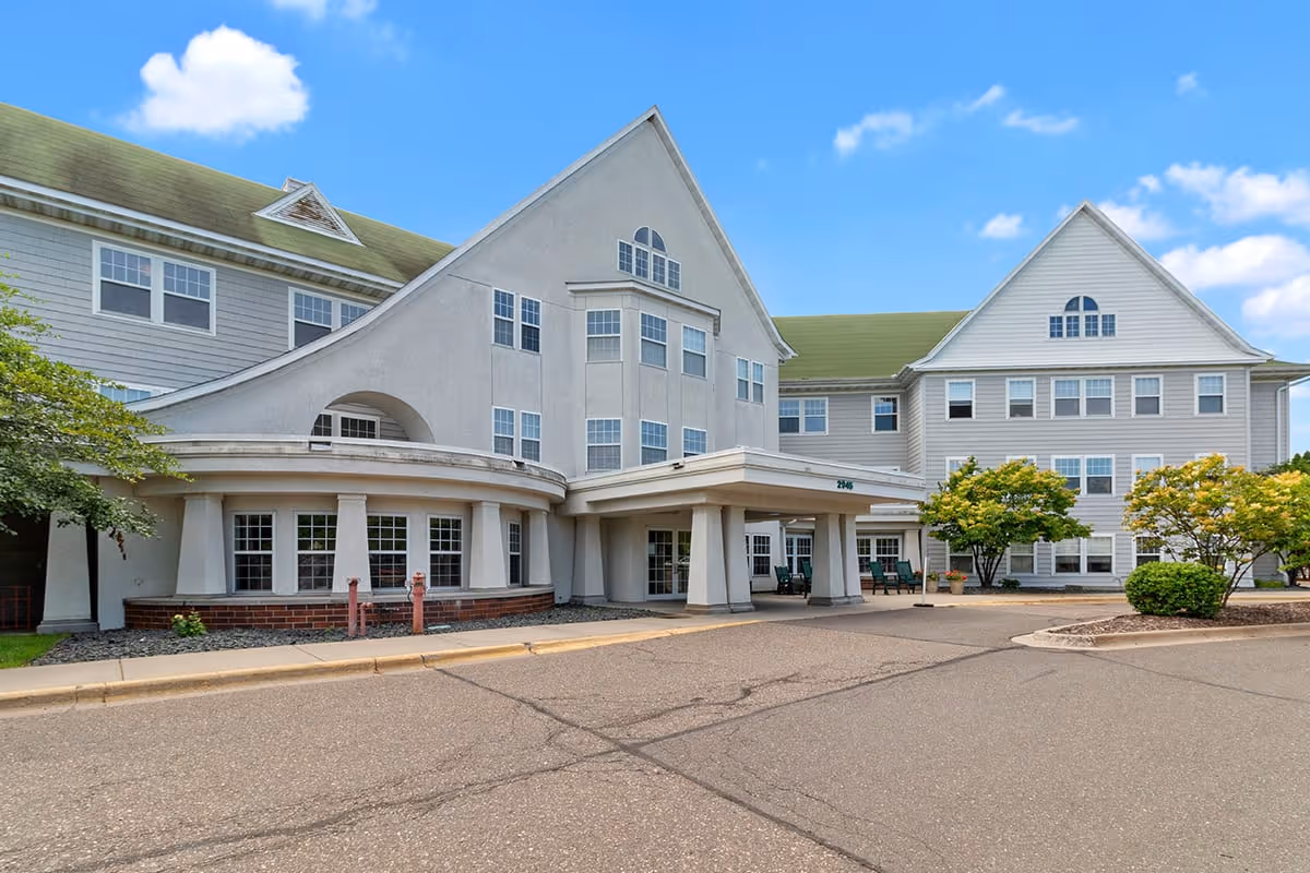 Exterior view of Eagle Crest Assisted Living facility showing a large multi-story building with white siding and green roof. The entrance has a covered drop-off area with columns, surrounded by trees and shrubs under a blue sky with some clouds.