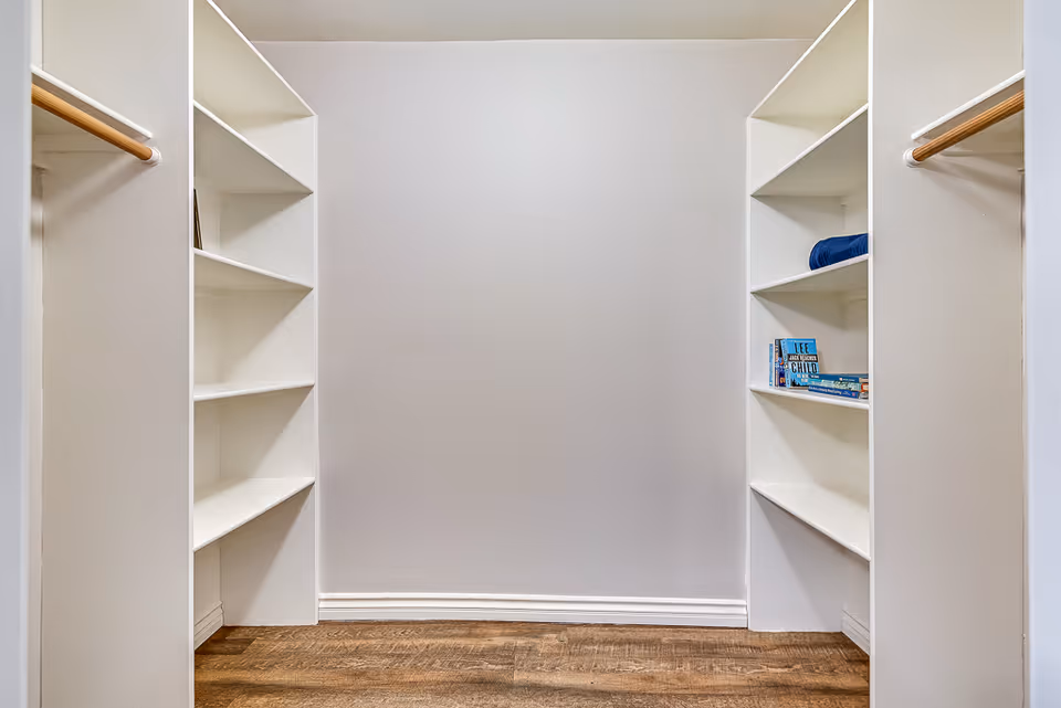 Empty walk-in closet with white built-in shelves on both sides and wooden hanging rods. A few books and a folded blue towel are placed on the shelves on the right side. The floor is wooden and the walls are painted light gray.
