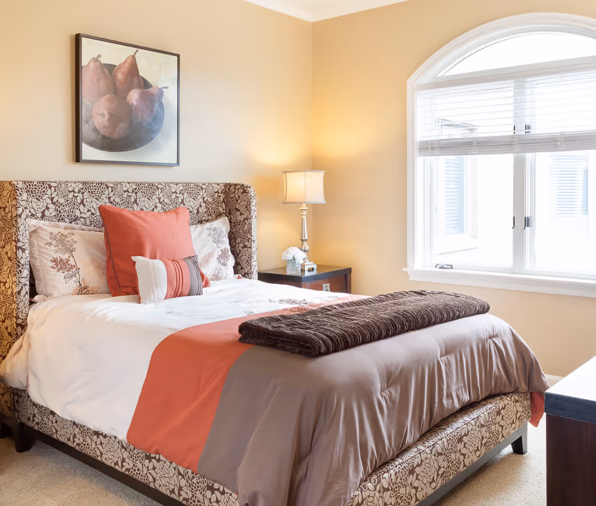 A cozy bedroom with a patterned upholstered bed frame, white and floral bedding with coral and brown accents, a folded brown blanket at the foot of the bed, a bedside table with a lamp and a small flower arrangement, and a large arched window with white blinds letting in natural light.
