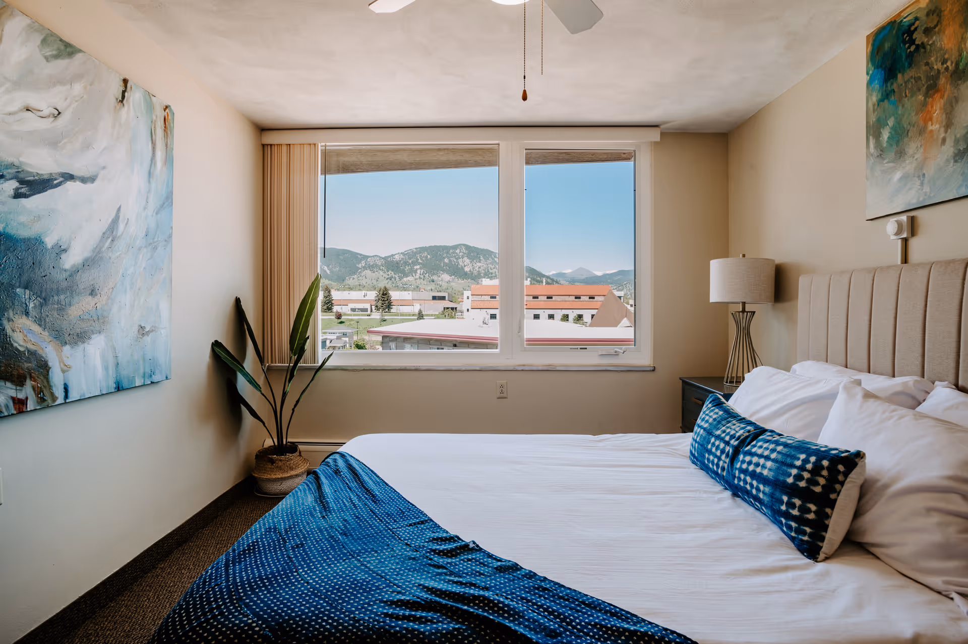 A bedroom with a large window showing a view of buildings and mountains in the distance. The room features a bed with white sheets and a blue patterned throw blanket and pillow. There is a bedside table with a lamp and a large abstract painting on the wall. A potted plant is placed near the window.