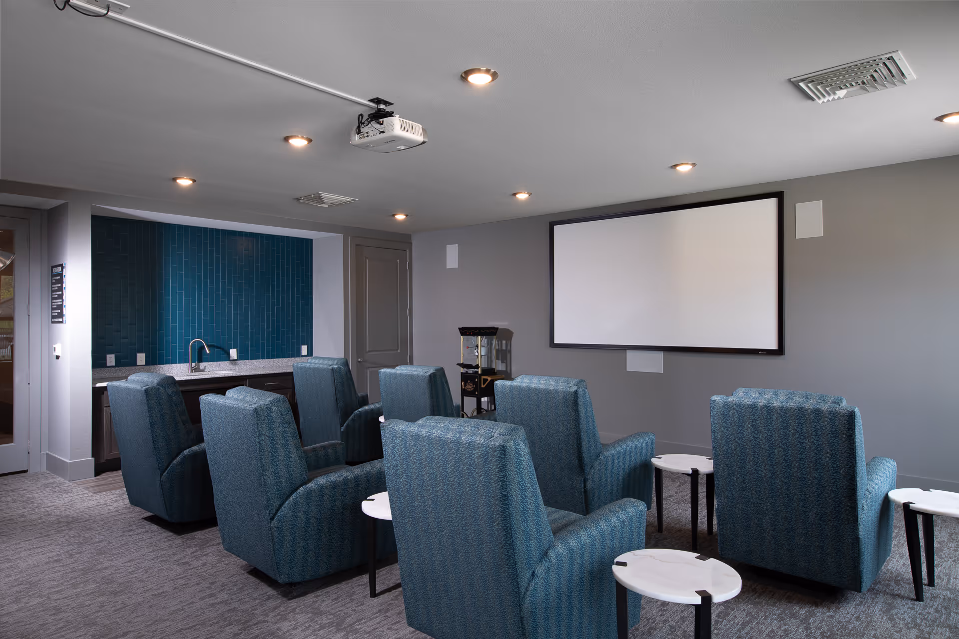 A small theater room with six blue upholstered recliner chairs arranged in two rows facing a large blank projector screen on a gray wall. There are small round white side tables next to the chairs. The room has a gray carpet, recessed ceiling lights, a ceiling-mounted projector, and a kitchenette area with a sink and blue tiled backsplash in the back corner.
