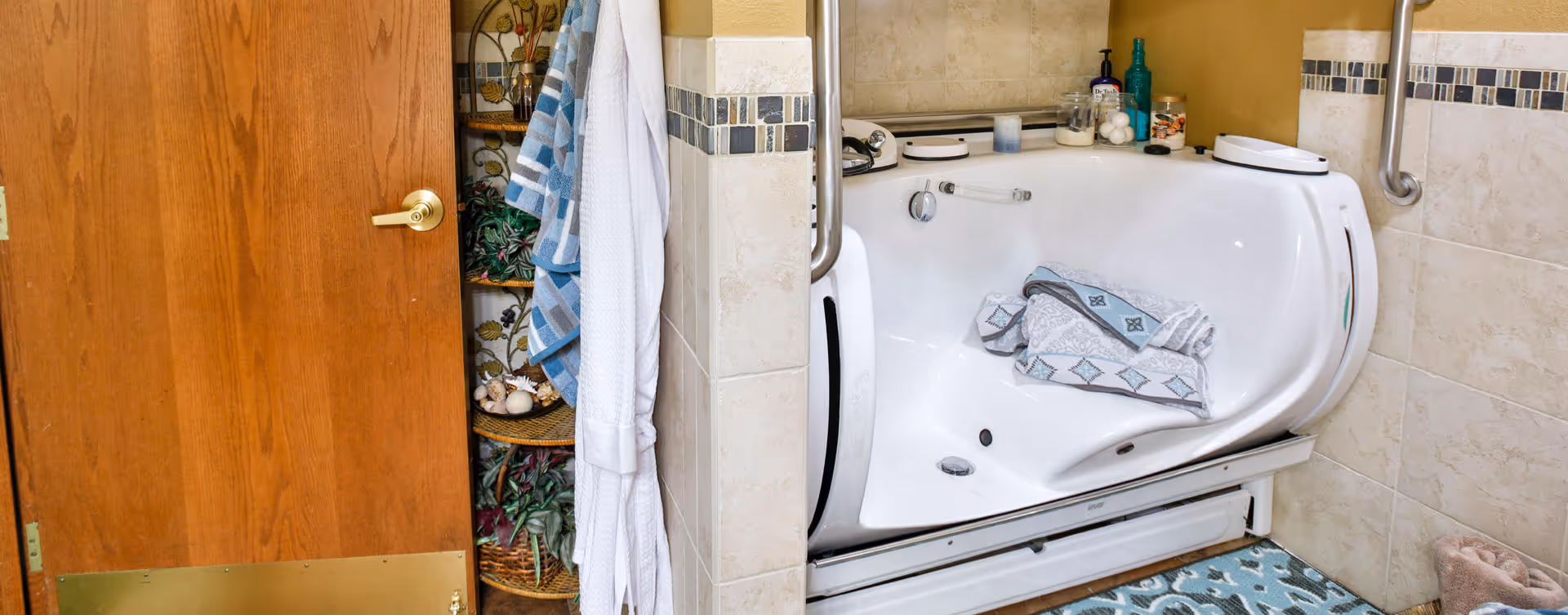 A bathroom featuring a white walk-in bathtub with a folded towel inside. The tub is surrounded by beige tiled walls with a decorative tile border. To the left, there is a wooden door with a brass handle and a white bathrobe hanging on a hook. A patterned blue and white rug is on the floor in front of the tub, and various bath products are placed on the tub's edge.