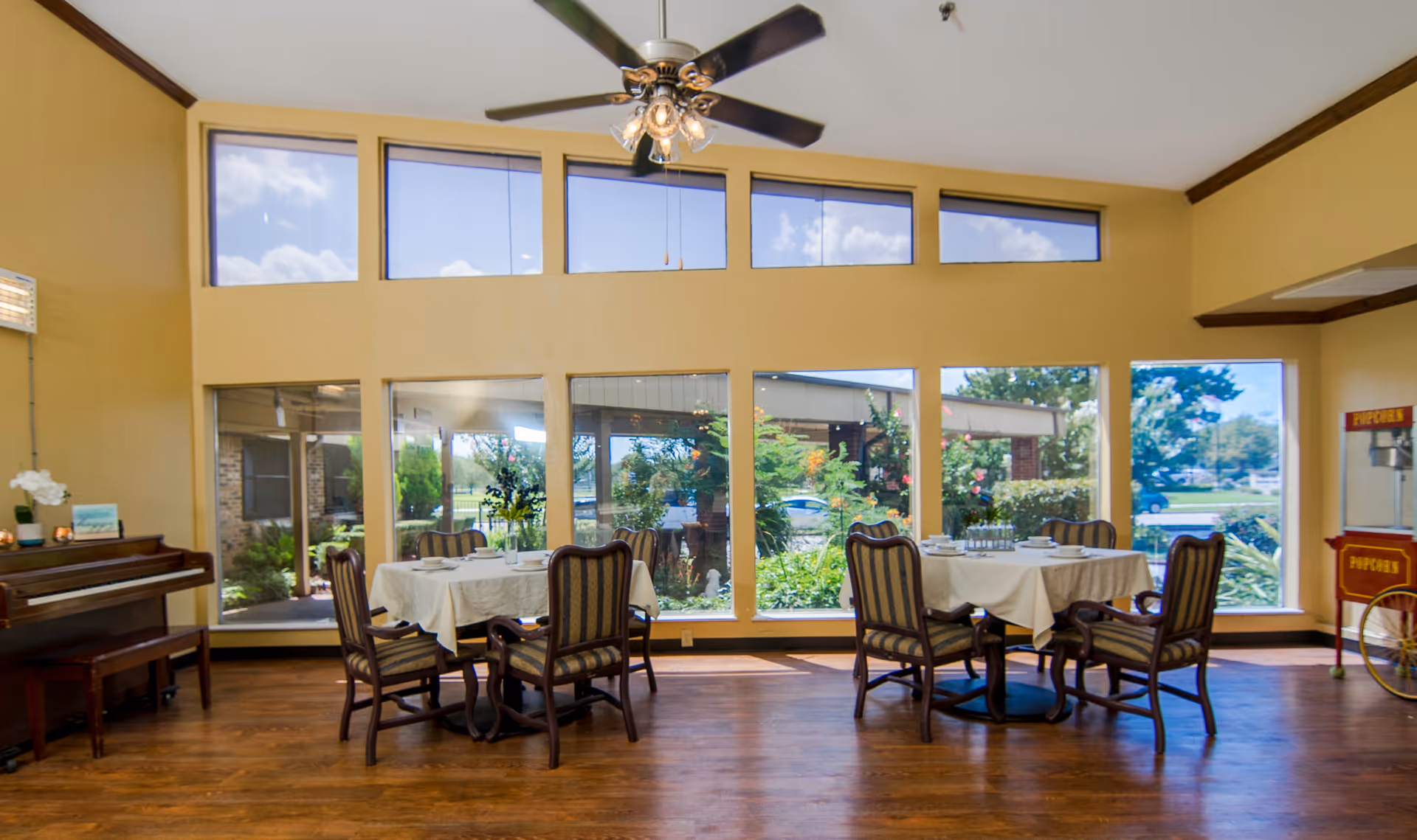Bright communal dining area with round tables and chairs by large windows overlooking a garden.