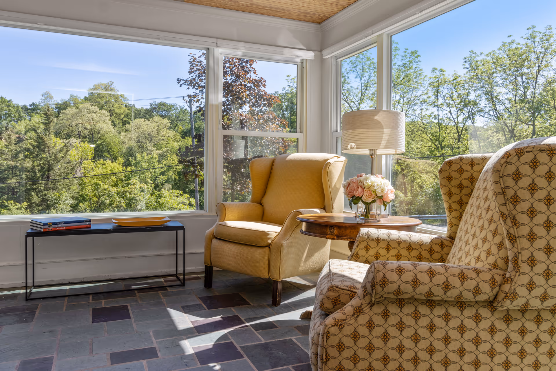 A bright sunroom with large windows showing a view of green trees outside. Inside, there are two patterned armchairs and one solid yellow armchair around a wooden side table with a lamp and a vase of flowers. A small black table with books and a plate is placed against the window.