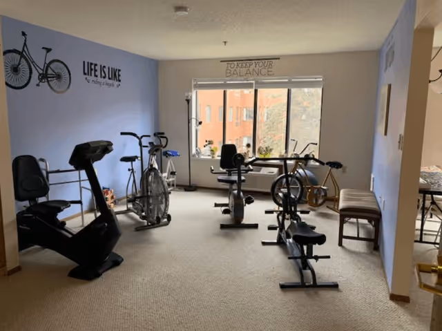 A fitness room with several exercise bikes and a rowing machine. The walls are painted light blue and white, with motivational quotes on the walls including a bicycle graphic and the phrase 'LIFE IS LIKE riding a bicycle' on the left wall and 'TO KEEP YOUR BALANCE' above the window. There is a large window letting in natural light and a bench along the right wall.