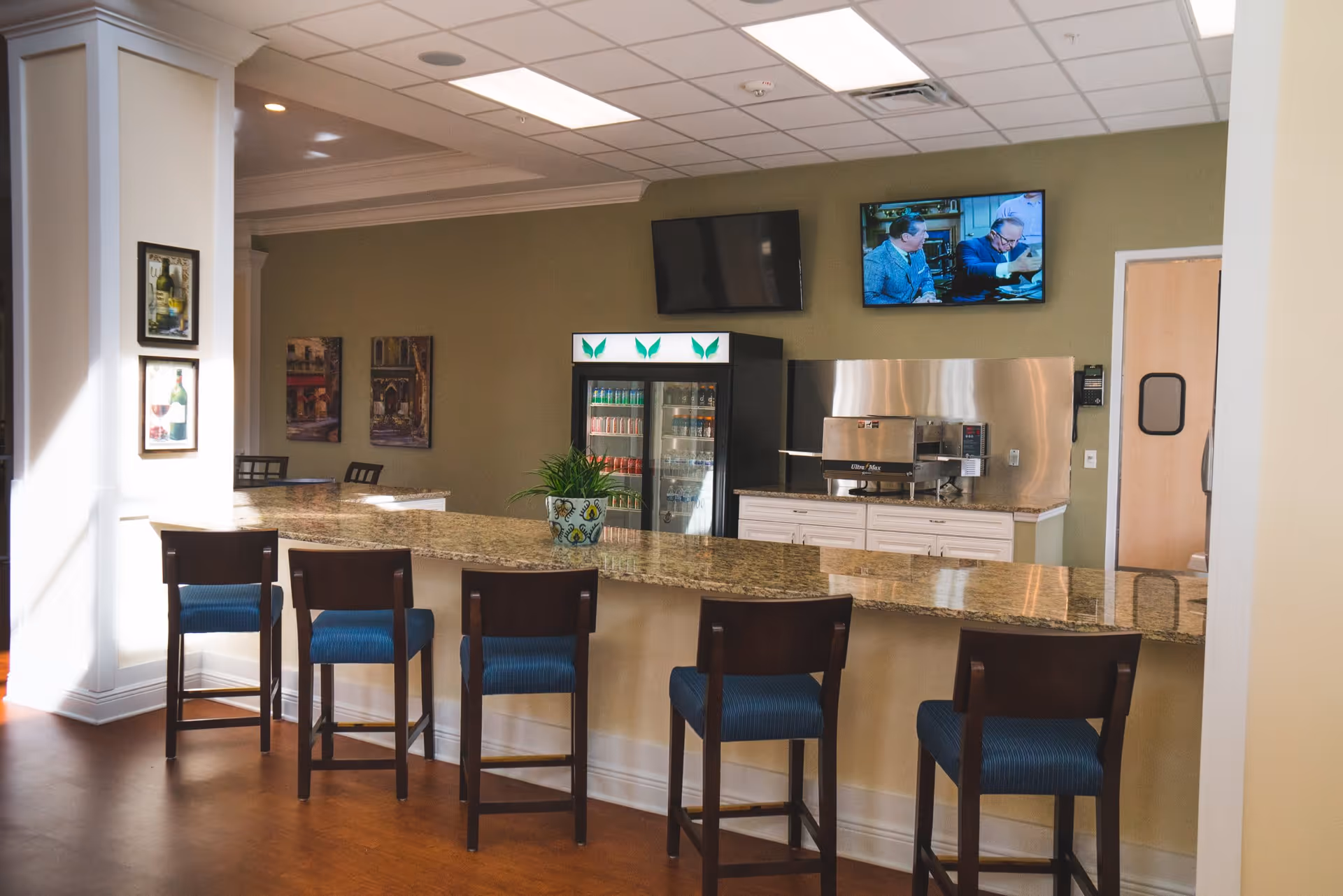 Interior view of a senior living facility's kitchen or snack bar area with a granite countertop and five wooden chairs with blue cushions. Behind the counter, there is a beverage refrigerator stocked with drinks, a coffee machine, and two wall-mounted televisions, one showing a scene from a movie or TV show. The walls are painted green and decorated with framed pictures.