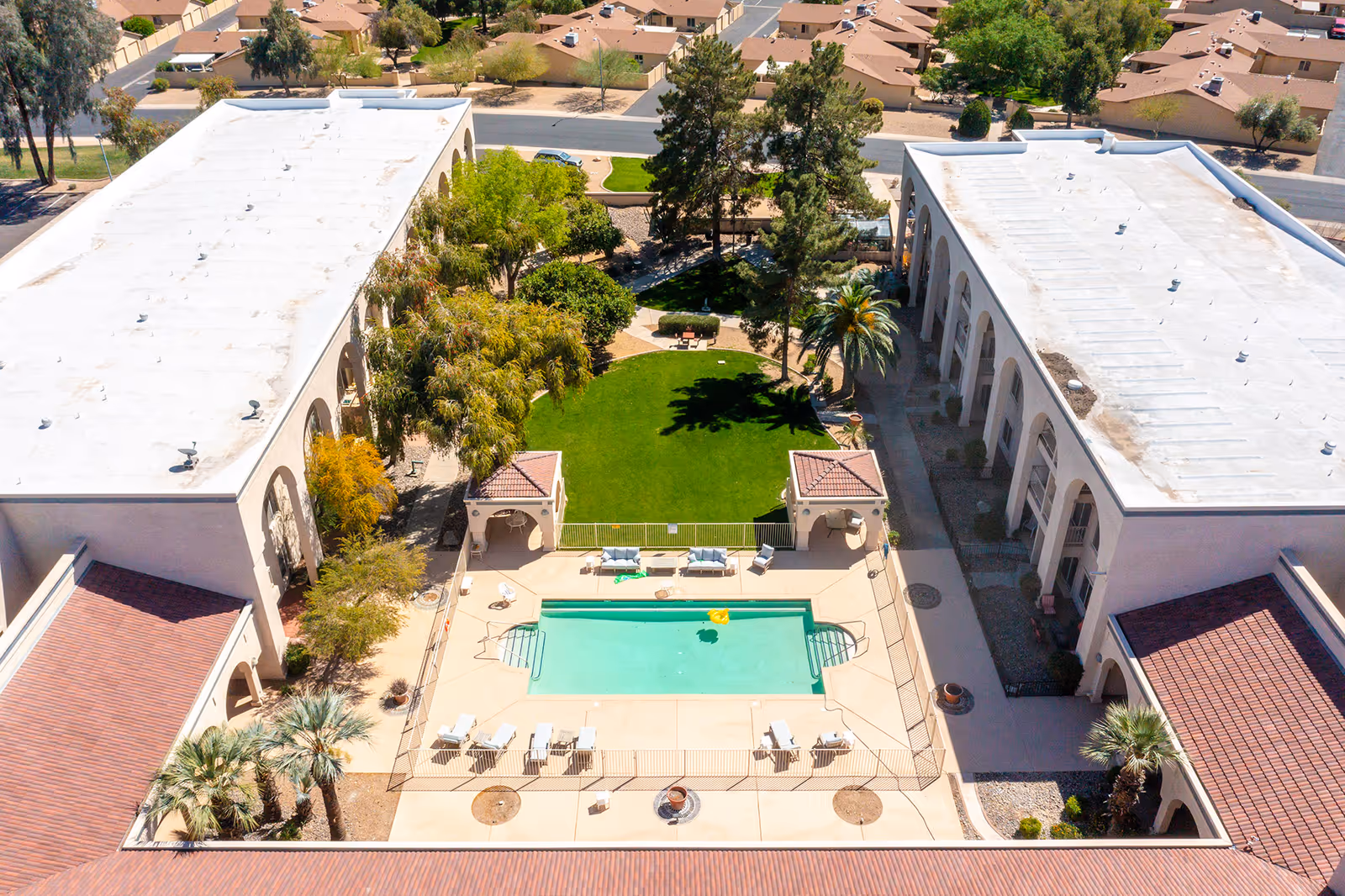 Aerial view of a senior living facility courtyard featuring a rectangular swimming pool surrounded by lounge chairs and two small covered seating areas. The courtyard also includes a well-maintained green lawn with trees and benches, flanked by two long buildings with white roofs and arched walkways. Residential houses and streets are visible in the background.