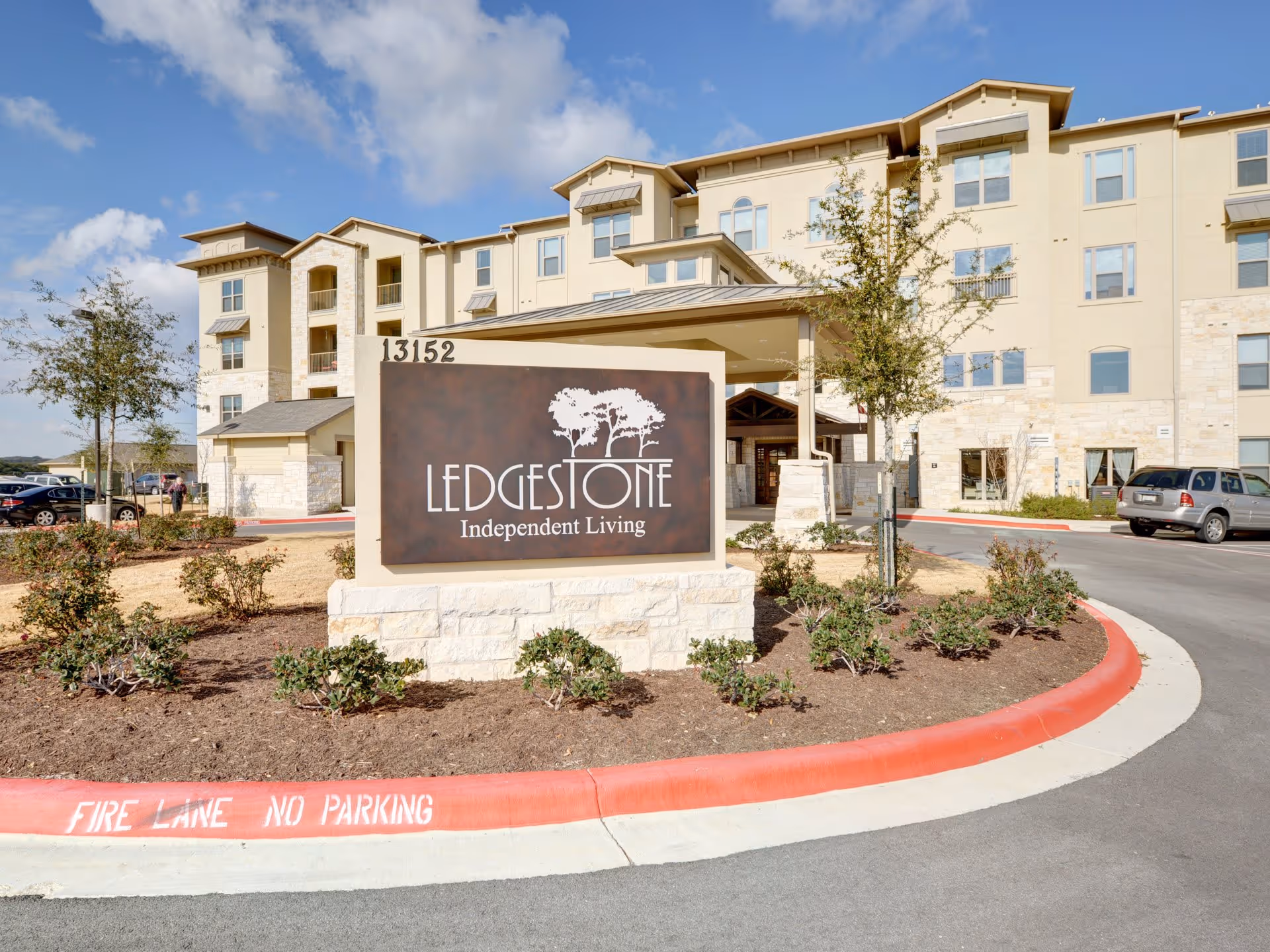 Exterior view of Ledgestone Senior Living building with a large sign in front that reads 'Ledgestone Independent Living'. The building is multi-story with beige and stone facade, surrounded by a landscaped area with small bushes and trees. A red curb with 'FIRE LANE NO PARKING' painted on it is visible in the foreground.