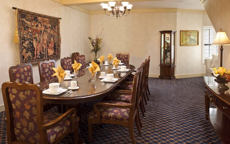 A formal dining room with a long wooden table set for a meal. The table has white cups, plates, silverware, and yellow folded napkins. The chairs around the table have wooden frames and purple upholstery with a leaf pattern. The room features a tapestry on one wall, a glass display cabinet, a lamp on a side table, and a carpet with a blue and brown pattern.
