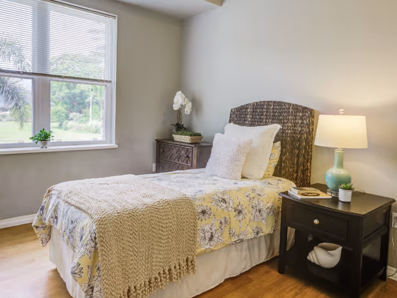 A cozy bedroom with a single bed featuring a floral yellow and white bedspread, a knitted beige throw blanket, and multiple pillows. Next to the bed is a dark wooden nightstand with a green lamp, a small plant, and a book. A wooden dresser with a white orchid plant is positioned against the wall. A large window with blinds lets in natural light, showing greenery outside.