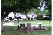 Outdoor area with picnic tables and benches on a grassy lawn, surrounded by trees and bushes. In the background, there are parked vehicles and residential-style buildings.