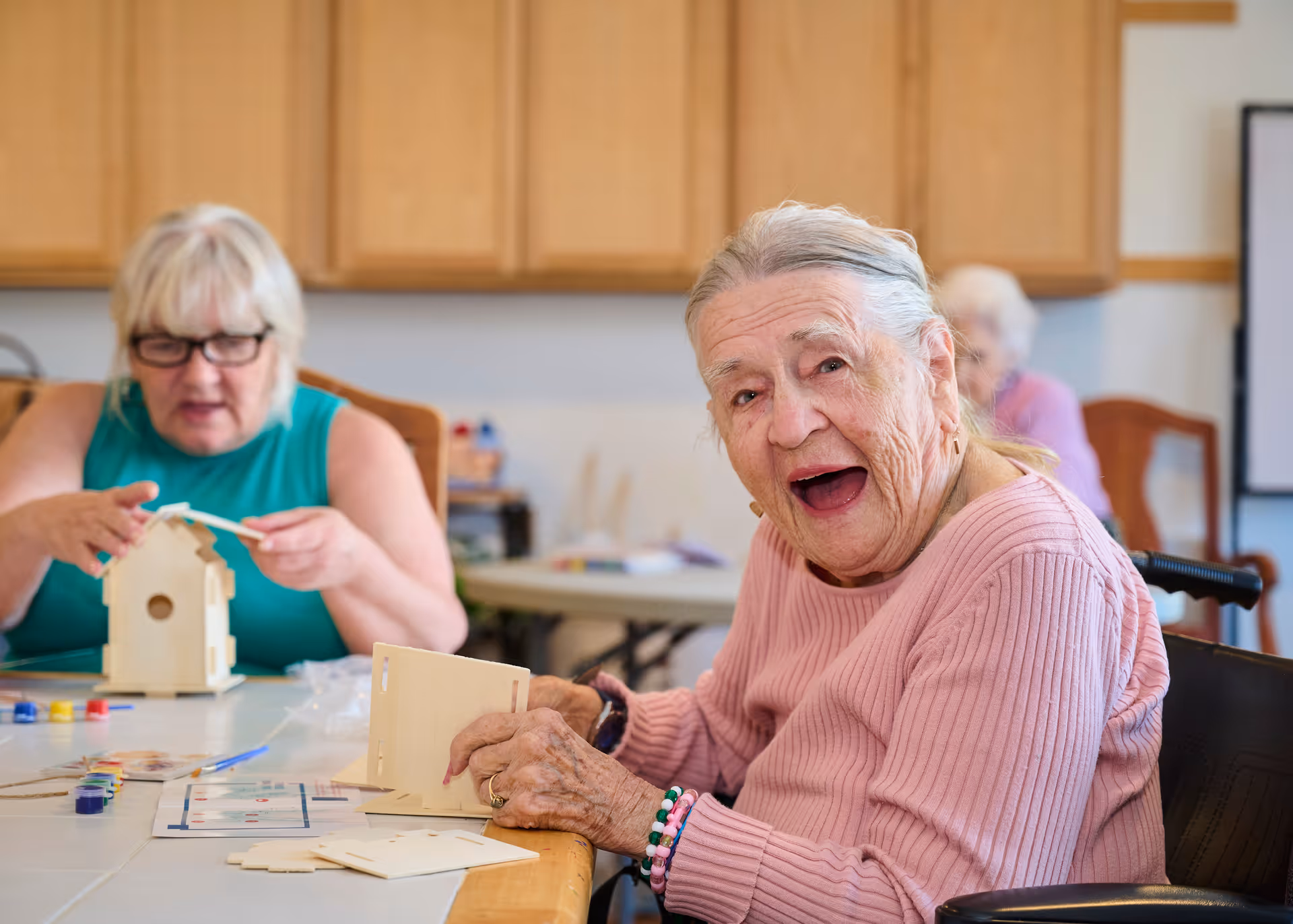 Two elderly women sitting at a table working on assembling and painting wooden birdhouses in a bright room with wooden cabinets in the background. One woman in a pink sweater is smiling and looking towards the camera, while the other woman in a teal sleeveless top is focused on her birdhouse.