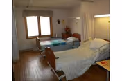 Two hospital-style beds in a shared senior care room with a window, privacy curtain, and wood flooring.